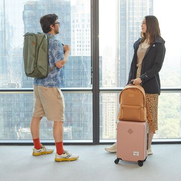 best travel backpack - index two people in front of windows one is carrying a large green backpack and another has a light tan backpack on top of a pink carry on luggage