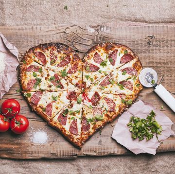 Heart-shaped pizza sliced heart shaped pizza with toppings on a wood cutting booard with tomatoes, basil, shredded cheese and pizza cutter