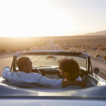best road trip songs   rear view of couple on road trip driving classic convertible car towards sunset